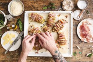 Sfornare e inserire tra le fessure delle patate del formaggio Emmentaler tagliato a fettine sottili e il prosciutto di San Daniele