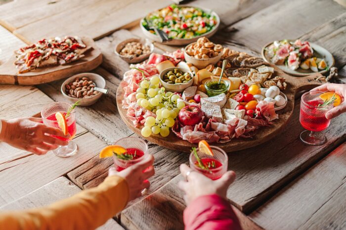 tagliere con salumi, frutta fresca e formaggi e persone che festeggiano con un cocktail in mano