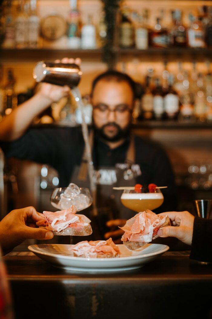barman che prepara dei cocktail mentre i clienti mangiano un crostino di pane con una fetta di prosciutto di san daniele