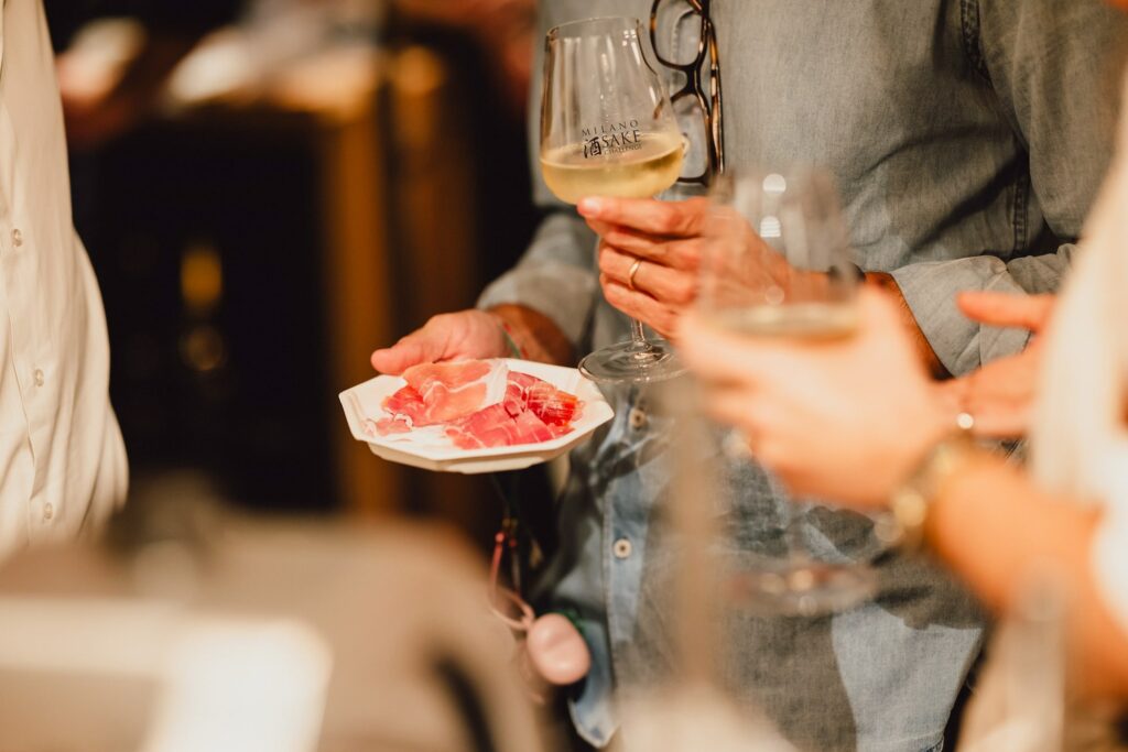 persona con un calice di saké e un piatto con fette di prosciutto di San Daniele in abbinamento