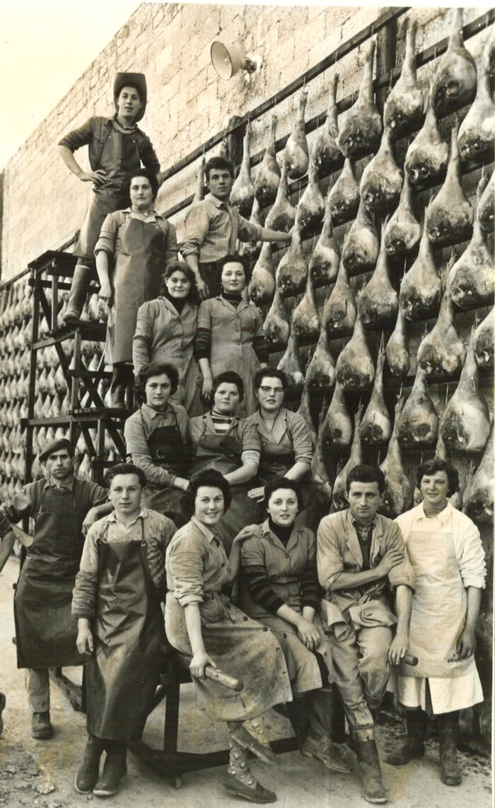 foto storica di gruppo con persone che posano davanti a una parete ricoperta di cosce di Prosciutto di San Daniele