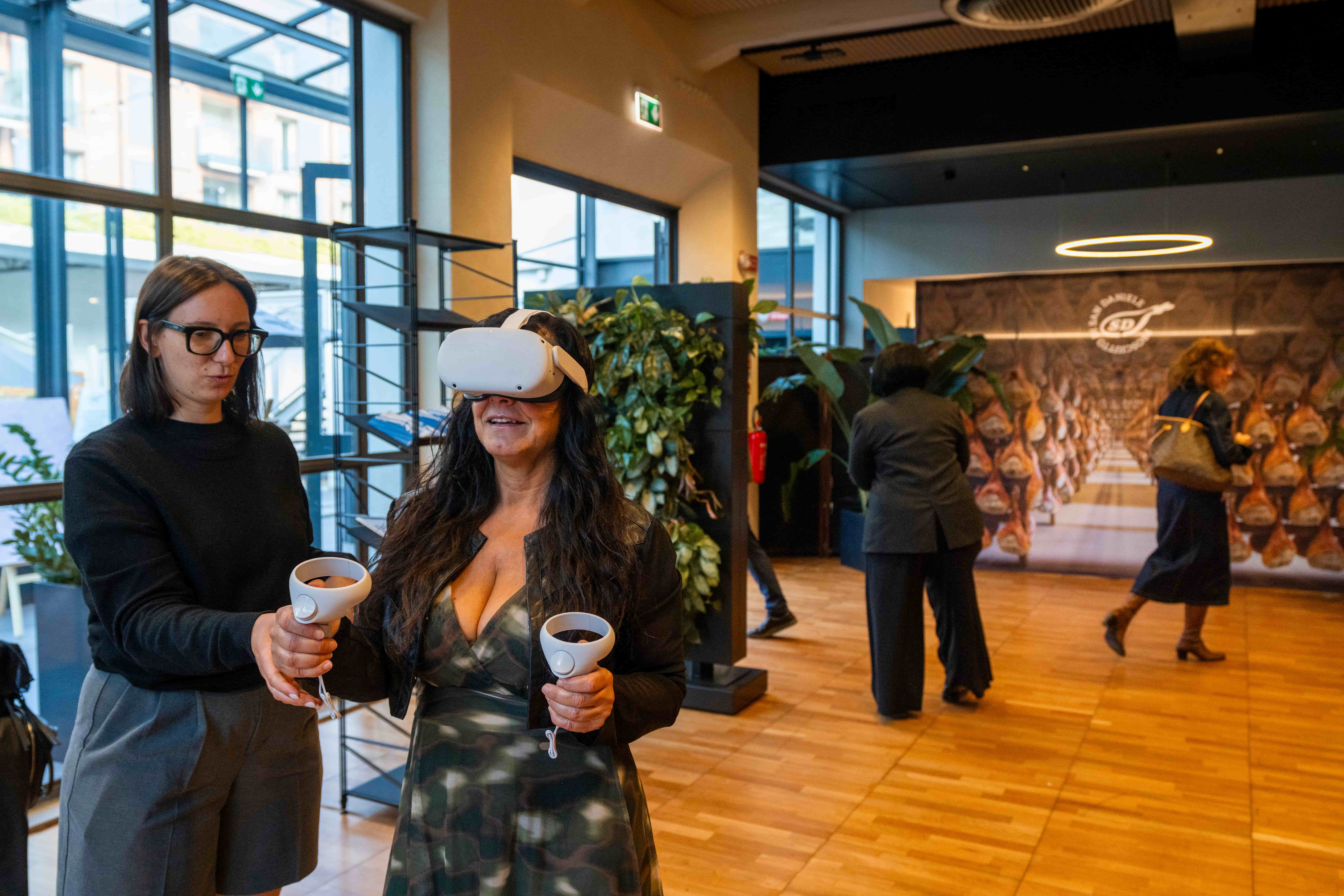 Donna sorridente indossa un visore VR Meta Quest e impugna i controller durante una dimostrazione interattiva, affiancata da un'assistente, in uno showroom dedicato al prosciutto San Daniele con stanze di stagionatura sullo sfondo.