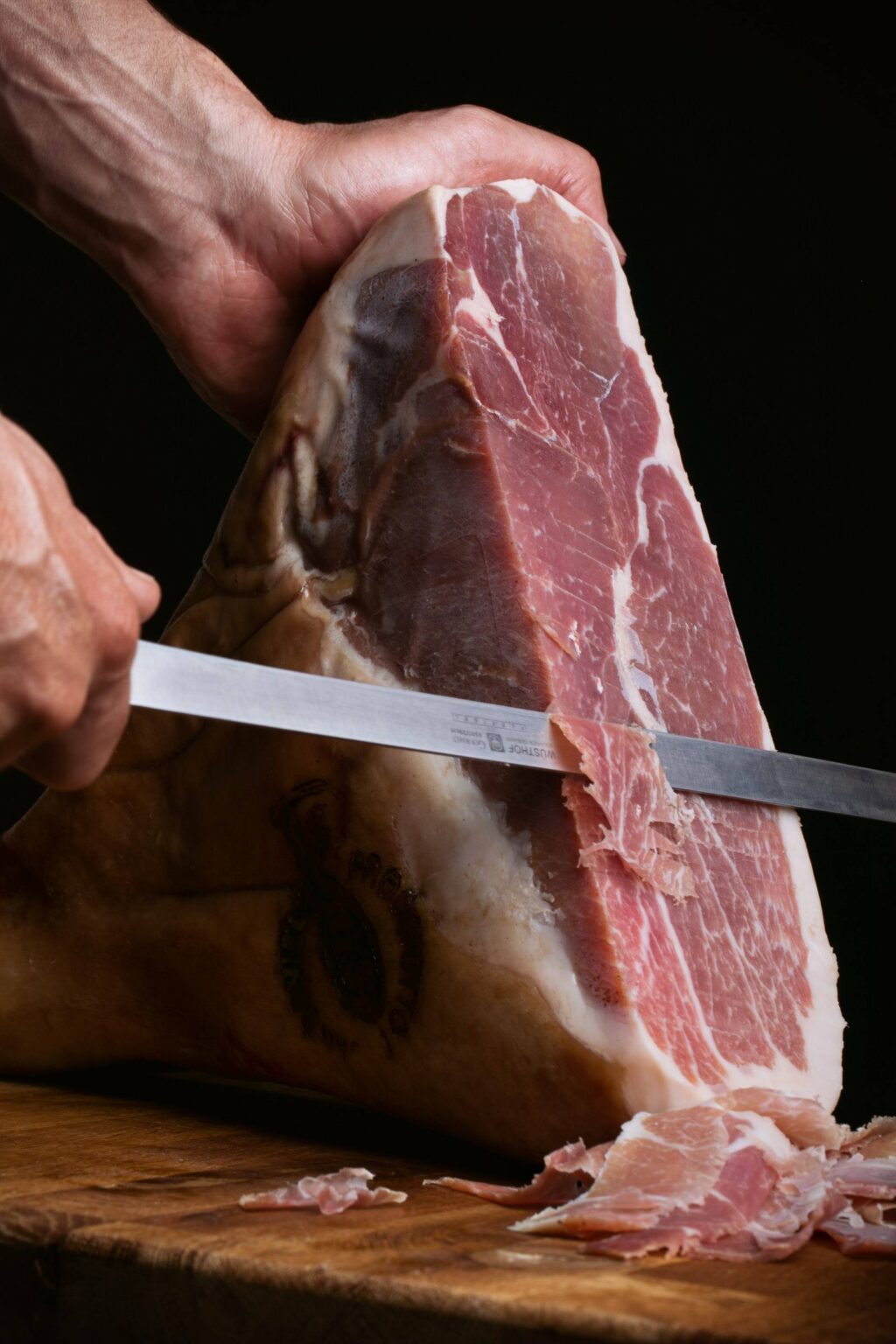 Mani che affettano al coltello un Prosciutto di San Daniele DOP su tagliere di legno, con il marchio a fuoco visibile sulla cotenna